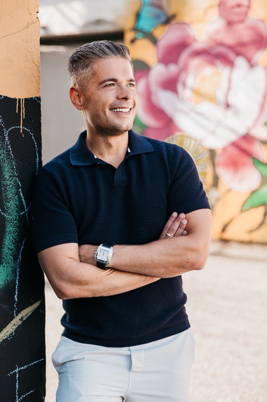 Andrew James Gray, arms crossed, smiling in front of a colourful Vancouver mural